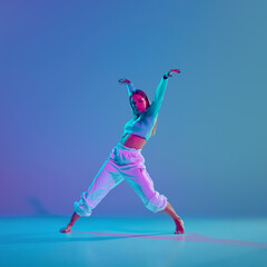 Youthful energy, dynamic hip hop choreography. Young talented and artistic girl performing against gradient blue background in neon light. Concept of freestyle dance, modernity, urban culture, youth