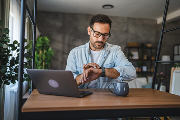 Adult man check time on smart watch while working from home on laptop