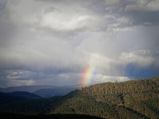 Landscape of mountains covered with trees under a storm, with the rainbow peeking through the clouds