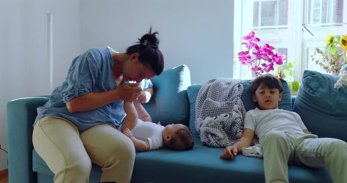 Mother interacting with her baby while sitting on the sofa with her other child, creating a warm and loving family moment in a cozy, light-filled living room