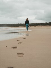 Woman walking barefoot on a sandy beach leaving footprints behind on a cloudy day.