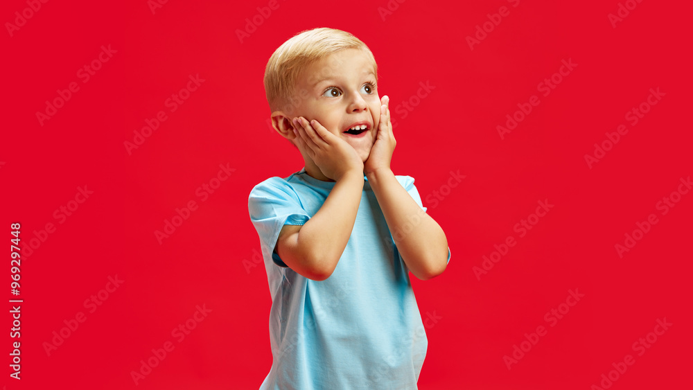 Wall mural Portrait of little blonde-haired boy, child in blue t-shirt expressing positive excitement, holding hands to face against red studio background. Concept of childhood, emotions, fashion, lifestyle