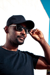 Afro black guy on the city center wearing black clothes cap an sun glasses modeling touching sunglasses with a white background.