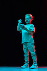 Little blonde-haired boy in blue t-shirt scrolling mobile phone against black studio background in neon light. Online cartoons, games. Concept of childhood, emotions, fashion, lifestyle