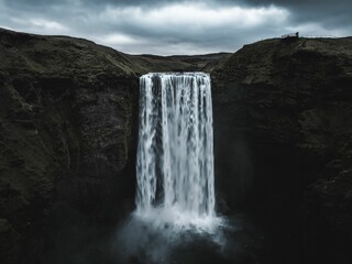 Waterfall cascades from a rocky cliff under an overcast sky, captured by drone in Iceland