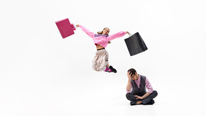 Woman leaps into air, filled with shopping joy, while her partner sits down in exhaustion against white studio background. Concept of shopping, Black Friday, fashion and style, sale season. Ad
