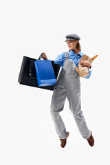 Dressed in casual overalls and cap, woman balances grocery and shopping bags in playful pose against white studio background. Concept of shopping, Black Friday, fashion and style, sale season. Ad