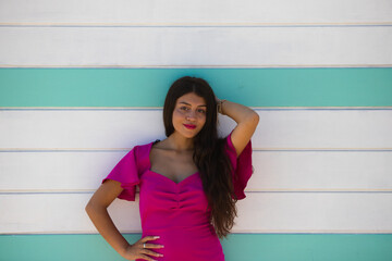 beautiful young woman with long hair in a pink dress, on the white and turquoise striped wooden wall of the beach changing rooms. The woman is on holiday in andalusia, Spain. Beauty and fashion.