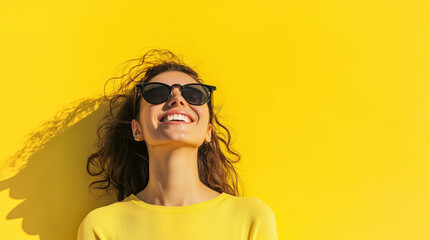 Fototapeta premium A joyful young woman with sunglasses, smiling happily while standing against a bold yellow wall, radiating positivity on a bright summer day. photo
