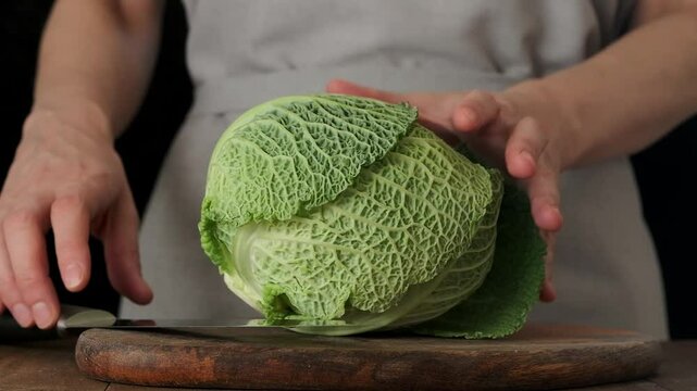 Female hands take a knife and cut head of Savoy cabbage in half on wooden chopping board. Slow motion.