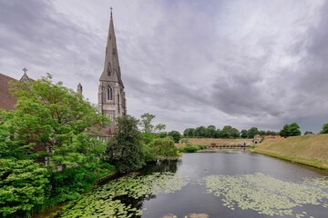 Fototapeta premium Church with tall spire next to pond under cloudy sky.