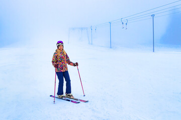 A young skier skillfully navigates snowy slopes in a vibrant outfit, surrounded by mist and winter scenery