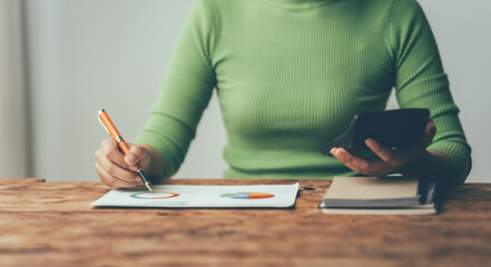 Focused on the Numbers: A woman meticulously analyzes charts and figures, her hand holding a calculator, reflecting the dedication and precision required for financial success. The image evokes a sens © Kritdanai
