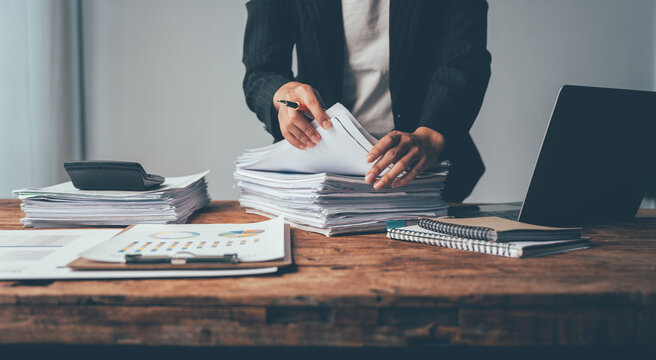 Overwhelmed by Paperwork: A businesswoman grapples with a towering stack of documents, her desk cluttered with the tools of a demanding profession, evoking the stress of deadlines and administrative o
