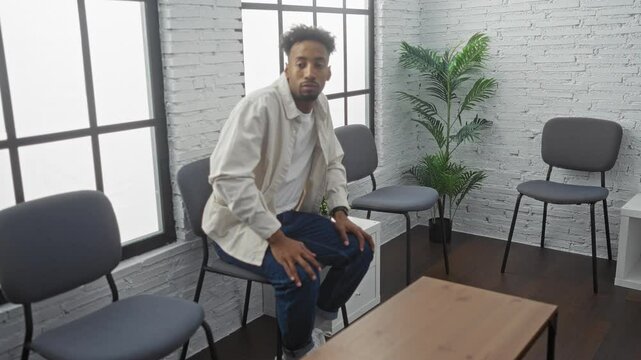 Young african american man waiting in an indoor room, checking time on his wristwatch, seated on a chair with modern decor and light filtering through large windows.