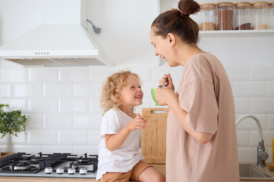 Happy mom sipping coffee and having fun with her cute child enjoying healthy drink together in the cozy kitchen and sharing smiles laughing while drinking tea in the morning
