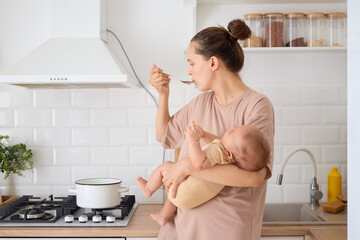 Caucasian woman with bun hairstyle mother standing in the kitchen multitasking with her newborn...