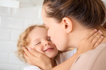 Beautiful mother hugging her adorable toddler kissing her satisfied daughter with eyes closed on the cheek enjoying tender moment of love and happiness