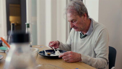 Elderly man eating alone at a dining table, concentrating on his meal. The scene conveys solitude and a quiet moment in a homey setting
