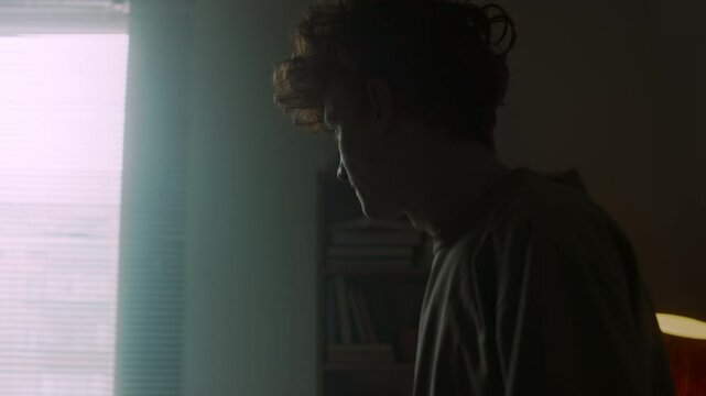 Hesitant young man looking at phone, then sitting on unmade bed and tossing gadget aside in reflection
