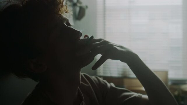 Close-up view of depressed young man sitting, leaning on wall with his head and smoking cigarette in bedroom against window