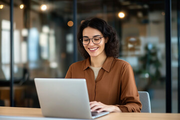 A smiling businesswoman in glasses uses a laptop in an office setting.