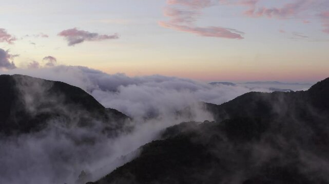 Aerial descending through wispy clouds towards mountain forest landscape and with pink and blue soft dawn sunrise in the background