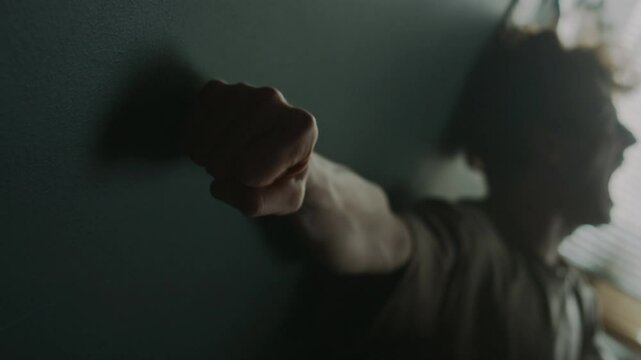 Rack focus shot of young man screaming and pounding dark grey wall with edge of palm clenched in fist in despair