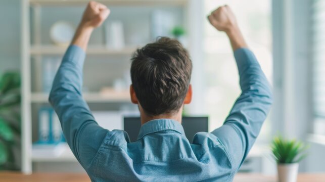 Rear view of a man in a blue shirt sitting at his desk, raising his arms in celebration, expressing excitement in a bright office setting.