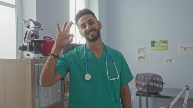 Young hispanic man in green medical scrubs making an ok gesture with his hand in a veterinary clinic, smiling and standing beside a microscope inside an indoor workplace.
