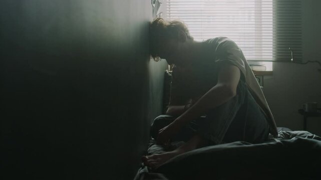 Frustrated young man rubbing his shoulder and forearm, then hitting head against grey wall and punching it with fist while sitting on unmade bed