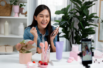 Young beautiful Asian reviewing cosmetic makeup and skin care collection, talking customer on social media online, presenting product surrounded beauty merchandise recording blurred phone . Stratagem.