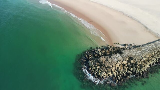 Ocean with empty sandy beach and stone breakwaters on a sunny day in Trafaria, Lisbon, Portugal