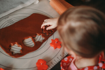 A child presses a gingerbread man-shaped cutter into brown dough on a baking mat. Various red...