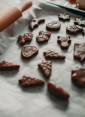 Raw gingerbread cookies in festive shapes like stars, trees, hearts, and gingerbread men on parchment paper, with a wooden rolling pin nearby.