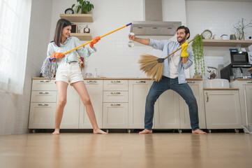 Happy couple having fun while doing cleaning kitchen, They use broom and mop instead of musical...