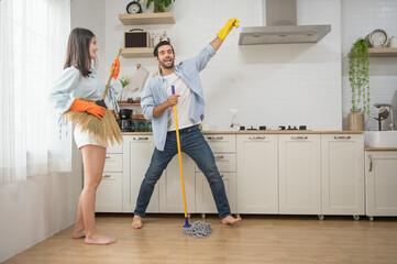 Happy couple having fun while doing cleaning kitchen, They use broom and mop instead of musical...