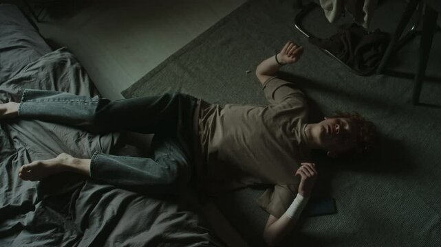Top down shot of exhausted young man lying on floor on his back with legs thrown on unmade bed holding phone, then turning on side, white bandage wrapped around his wrist