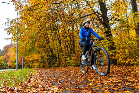 Middle aged woman wearing blue sporty jacket, black cycling pants and white bike helmet riding bicycle on bicycle path in city by forest in autumn scenery. Front view.	
