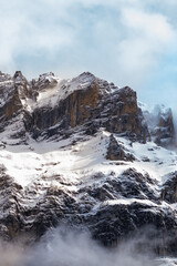 Vertical landscape  of a snowy mountain peak among the Alps in Grindelwald, Switzerland. Snow covers the rocky face of the mountain, and the sky is blue above with soft white clouds.