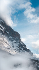 A snowy mountain among the Alps in Grindelwald, Switzerland. Snow covers the rocky face of the mountain, and the sky is blue above with soft white clouds. The mountain emerges from low clouds and mist
