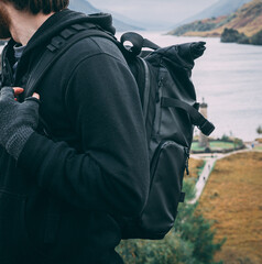 Crop of a male hiker and his backpack as he stands near some hills at Glenfinnan Monument in the Highlands, Scotland. He wears a non-branded camera backpack, black hoodie and fingerless gloves.
