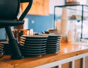 Stack of dishes on the counter of a cafe. They're next to an espresso machine. Coffee cups are in the background.
