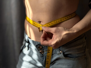 Close up of faceless woman in jeans measuring her waist with yellow tape measure. Weight loss, fitness and body measurement concept for health and wellness. Lifestyle photo.