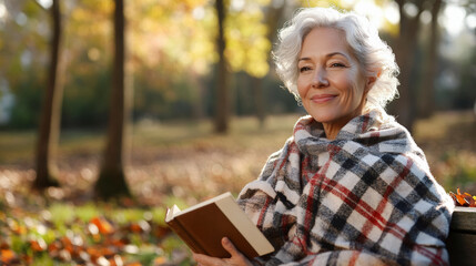 An elderly woman wrapped in a plaid blanket sits on a park bench, enjoying a peaceful moment reading a book during autumn.