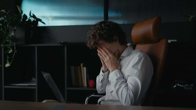 Side view of a tired guy with curly hair in a shirt getting tired and touching his face while working on a laptop in the office