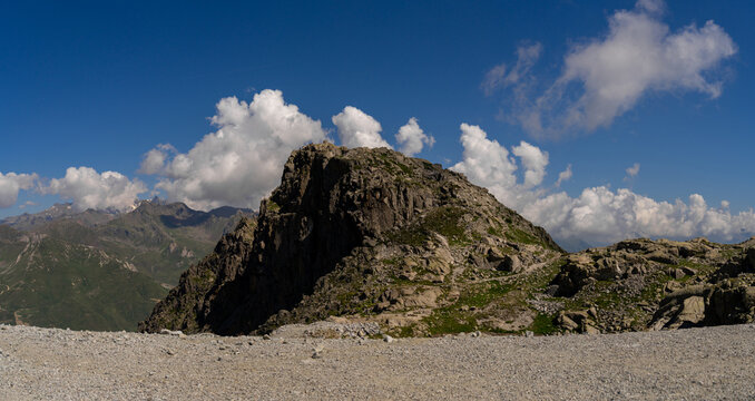 Panoramic view of rugged mountain peak set against clear blue sky, with scattered clouds. The rocky terrain in foreground contrasts with the distant green valleys, dramatic landscape