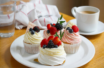 Cupcakes and mini pavlova cakes decorated with fresh berries and white chocolate on a large white plate, a cup of coffee, glass of water and a cloth napkin in the background. Selective focus.