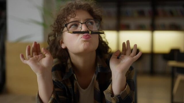 Funny girl student with curly hair wearing glasses puts a black pen on her upper lip and laughs while sitting at a table in the library