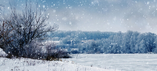 winter landscape with trees on the river bank and cloudy sky during snowfall